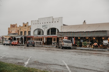 A small town street scene featuring historic buildings with old-fashioned architecture. The street is lined with several small businesses, including a bakery cafe and other shops, as indicated by colorful signs and awnings. Cars are parked along the street, and the overall atmosphere appears quiet and overcast.