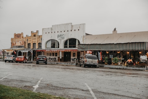 A small town street scene featuring historic buildings with old-fashioned architecture. The street is lined with several small businesses, including a bakery cafe and other shops, as indicated by colorful signs and awnings. Cars are parked along the street, and the overall atmosphere appears quiet and overcast.