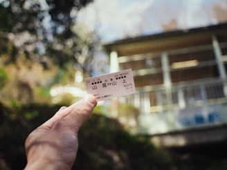 A hand holds a small ticket with Japanese writing against a blurred outdoor background featuring greenery and the side of a building.