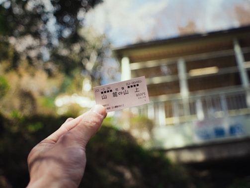 A hand holds a small ticket with Japanese writing against a blurred outdoor background featuring greenery and the side of a building.