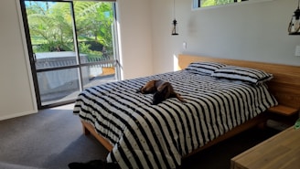 A bedroom with a minimalistic design featuring a bed with black and white striped bedding. A dog is resting in the middle of the bed. The room has a large sliding glass door leading to an outdoor area with lush green plants visible. Pendant lights hang from the ceiling, and the walls are painted a soft white.