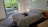 A bedroom with a minimalistic design featuring a bed with black and white striped bedding. A dog is resting in the middle of the bed. The room has a large sliding glass door leading to an outdoor area with lush green plants visible. Pendant lights hang from the ceiling, and the walls are painted a soft white.