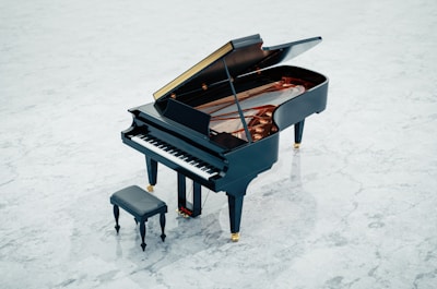 A grand piano with its lid open, showcasing the strings and internal components, placed on a light grey marble floor. There is a matching piano bench positioned in front of the piano.