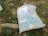 A large white sack with blue text lies on a patch of ground with scattered rocks and pieces of wood. The text appears to be in a foreign language, likely Chinese, and includes a logo with the words 'GLORIA FLOUR.' The sack rests in an outdoor setting with a mix of grass and dirt.