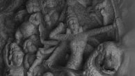 A detailed stone carving depicting a warrior goddess engaged in battle. The central figure, adorned in intricate garments, wields weapons with fierce determination, surrounded by other figures and mythical creatures. The artistry highlights dynamic motion and expressive detail.