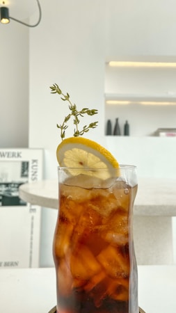 A tall glass filled with iced tea garnished with a slice of lemon and a sprig of thyme is placed on a white table. The background features a minimalist interior with shelves and decorative elements in soft tones.