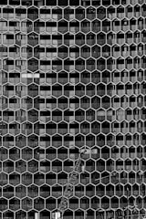An intricate facade featuring a hexagonal grid design covers a building, creating a geometric pattern. A construction crane is positioned in front of the structure, indicating ongoing development or maintenance. The image is black and white, highlighting the contrast between the architectural details and the crane.