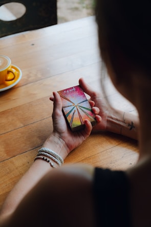A person holding a deck of tarot cards with a colorful and abstract design on the top card. The person wears bracelets and has a tattoo of a bird on their arm. A cup with a saucer is placed on the wooden table beside them.