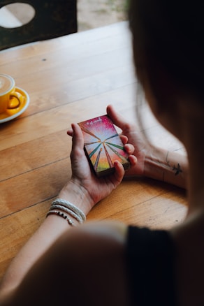 A person holding a deck of tarot cards with a colorful and abstract design on the top card. The person wears bracelets and has a tattoo of a bird on their arm. A cup with a saucer is placed on the wooden table beside them.