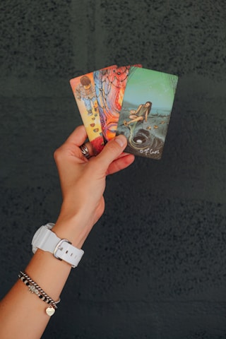 A hand wearing a white watch and bracelets is holding three tarot cards. The cards have colorful and detailed illustrations, including a child with cups, vibrant abstract patterns, and a person sitting with a cup near a hole.