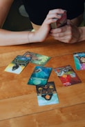 A wooden table with several tarot cards laid out, depicting various images and symbols. A person's hands are visible, holding a deck of cards ready for reading. The atmosphere suggests a moment of concentration and mystery.
