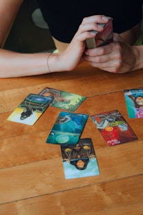 A wooden table with several tarot cards laid out, depicting various images and symbols. A person's hands are visible, holding a deck of cards ready for reading. The atmosphere suggests a moment of concentration and mystery.
