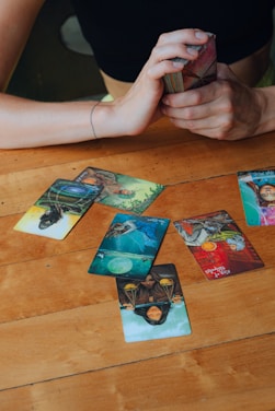 A wooden table with several tarot cards laid out, depicting various images and symbols. A person's hands are visible, holding a deck of cards ready for reading. The atmosphere suggests a moment of concentration and mystery.