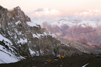 A rugged mountain landscape with peaks capped in snow stretches into the distance. The foreground features rocky terrain where two yellow tents are pitched, suggesting a campsite. The sky is painted with soft hues of pink and orange as the sun sets, casting a gentle glow on the mountain range.