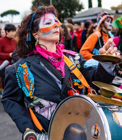 A person with colorful face paint and curly hair plays a drum in a lively outdoor setting. They wear a vibrant outfit with a dark jacket adorned with colorful patches and beads. A pink scarf is tied around their neck, and they hold a small cymbal in one hand. In the background, other people in similar festive attire are visible, suggesting a parade or celebration.