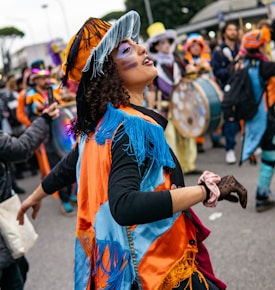 A person wearing vibrant and colorful attire, including a fringed orange and blue jacket and a matching hat, participates in a lively street parade. The scene is energetic, with other costumed individuals and musical instruments visible in the background.