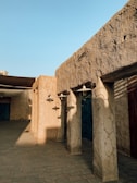 An architectural scene depicting traditional mud-brick structures with rustic textures. Shadows of outdoor lamps are cast on the walls, and wooden doors feature prominently. The lighting is warm, suggesting a late afternoon or early evening setting.
