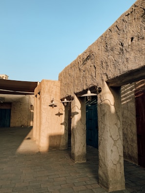 An architectural scene depicting traditional mud-brick structures with rustic textures. Shadows of outdoor lamps are cast on the walls, and wooden doors feature prominently. The lighting is warm, suggesting a late afternoon or early evening setting.