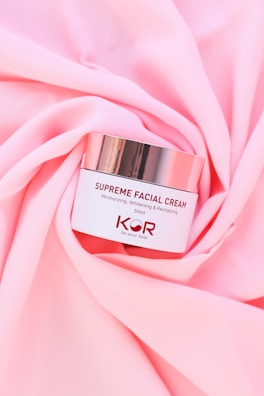 A jar of Supreme Facial Cream with a metallic lid is placed elegantly on soft, pink fabric. The cream is labeled as moisturizing, whitening, and revitalizing, with a confident tagline beneath the brand name.