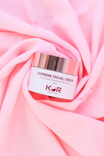 A jar of Supreme Facial Cream with a metallic lid is placed elegantly on soft, pink fabric. The cream is labeled as moisturizing, whitening, and revitalizing, with a confident tagline beneath the brand name.