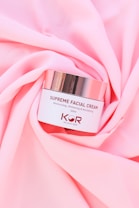 A jar of Supreme Facial Cream with a metallic lid is placed elegantly on soft, pink fabric. The cream is labeled as moisturizing, whitening, and revitalizing, with a confident tagline beneath the brand name.