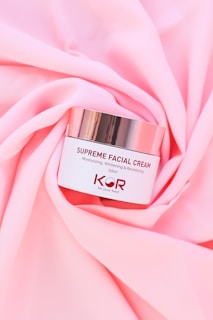 A jar of Supreme Facial Cream with a metallic lid is placed elegantly on soft, pink fabric. The cream is labeled as moisturizing, whitening, and revitalizing, with a confident tagline beneath the brand name.