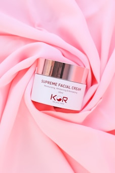 A jar of Supreme Facial Cream with a metallic lid is placed elegantly on soft, pink fabric. The cream is labeled as moisturizing, whitening, and revitalizing, with a confident tagline beneath the brand name.