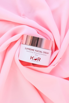 A jar of Supreme Facial Cream with a metallic lid is placed elegantly on soft, pink fabric. The cream is labeled as moisturizing, whitening, and revitalizing, with a confident tagline beneath the brand name.