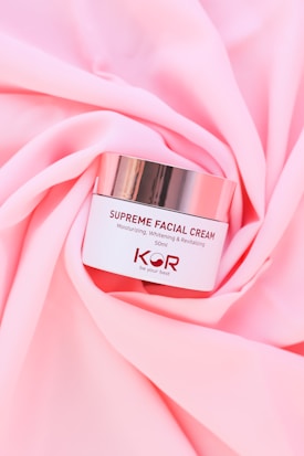 A jar of Supreme Facial Cream with a metallic lid is placed elegantly on soft, pink fabric. The cream is labeled as moisturizing, whitening, and revitalizing, with a confident tagline beneath the brand name.