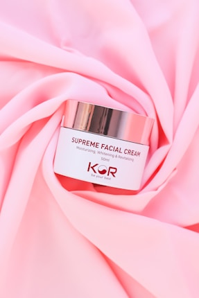 A jar of Supreme Facial Cream with a metallic lid is placed elegantly on soft, pink fabric. The cream is labeled as moisturizing, whitening, and revitalizing, with a confident tagline beneath the brand name.