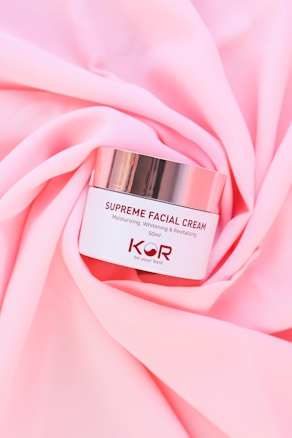 A jar of Supreme Facial Cream with a metallic lid is placed elegantly on soft, pink fabric. The cream is labeled as moisturizing, whitening, and revitalizing, with a confident tagline beneath the brand name.