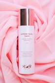 A bottle of Supreme Facial Toner with a cylindrical shape and a shiny metallic cap is placed on a soft, pink fabric backdrop. The label text describes its hydrating, refreshing, and smoothing properties. The design is minimalistic with the brand logo and instructions on the bottle.