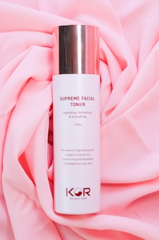 A bottle of Supreme Facial Toner with a cylindrical shape and a shiny metallic cap is placed on a soft, pink fabric backdrop. The label text describes its hydrating, refreshing, and smoothing properties. The design is minimalistic with the brand logo and instructions on the bottle.