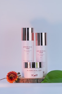A trio of facial skincare products, including a toner, serum, and cream, are neatly arranged on a wooden slab with a decorative flower and a green leaf beside them. The products have metallic caps, and the packaging features minimalist design with red and black text.