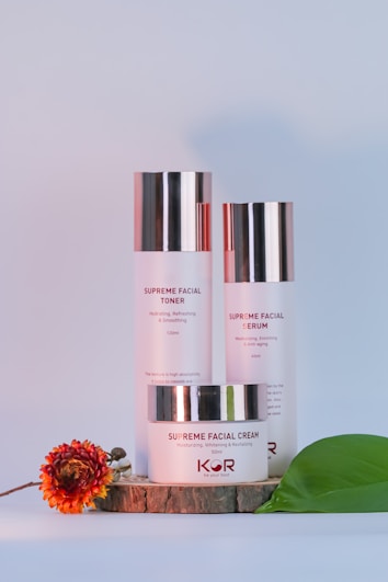 A trio of facial skincare products, including a toner, serum, and cream, are neatly arranged on a wooden slab with a decorative flower and a green leaf beside them. The products have metallic caps, and the packaging features minimalist design with red and black text.