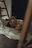 A person is lying on the floor, partially covered by a light-colored sheet in a dimly lit room. A wooden ladder is nearby, and there are hints of shadows and soft textures around. The mood is calm and introspective.
