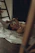 A person is lying on the floor, partially covered by a light-colored sheet in a dimly lit room. A wooden ladder is nearby, and there are hints of shadows and soft textures around. The mood is calm and introspective.