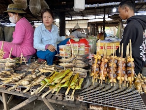 A market stall featuring a variety of grilled and roasted foods on skewers, including fish and possibly chicken. Two women and a man are present, with one woman wearing a mask. The stall is in a rustic, covered market area with various containers and equipment visible in the background.