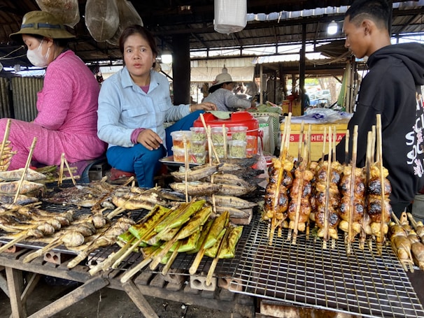 A market stall featuring a variety of grilled and roasted foods on skewers, including fish and possibly chicken. Two women and a man are present, with one woman wearing a mask. The stall is in a rustic, covered market area with various containers and equipment visible in the background.
