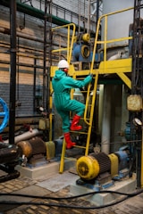 A person wearing a protective suit, hard hat, and red boots is climbing a yellow metal ladder inside an industrial facility. Various machinery components, including motors and pipes, are visible, with blue and yellow equipment prominently featured. The scene is set in a brick-walled, industrial environment with a complex network of metal structures and electrical equipment.