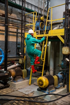 A person wearing a protective suit, hard hat, and red boots is climbing a yellow metal ladder inside an industrial facility. Various machinery components, including motors and pipes, are visible, with blue and yellow equipment prominently featured. The scene is set in a brick-walled, industrial environment with a complex network of metal structures and electrical equipment.