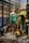 A person wearing a protective suit, hard hat, and red boots is climbing a yellow metal ladder inside an industrial facility. Various machinery components, including motors and pipes, are visible, with blue and yellow equipment prominently featured. The scene is set in a brick-walled, industrial environment with a complex network of metal structures and electrical equipment.