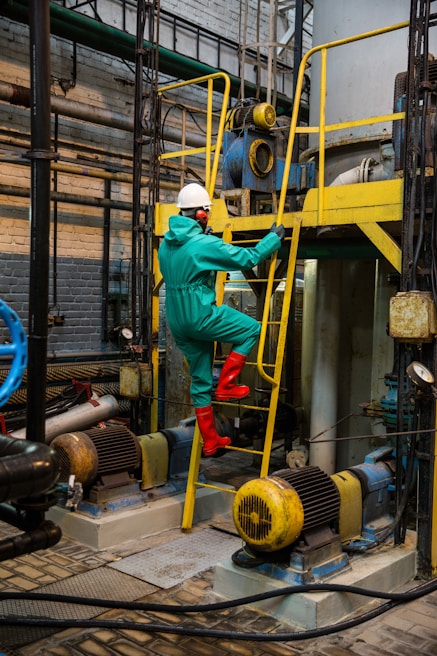 A person wearing a protective suit, hard hat, and red boots is climbing a yellow metal ladder inside an industrial facility. Various machinery components, including motors and pipes, are visible, with blue and yellow equipment prominently featured. The scene is set in a brick-walled, industrial environment with a complex network of metal structures and electrical equipment.