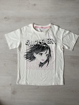 A white t-shirt with a printed graphic of a stylized cartoon girl's face, accompanied by bold black text saying 'ENJOY EVERY.' Below the graphic, the word 'MOMENT!' is repeated three times in light pink font. The shirt is laid flat on a light wooden surface.