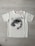 A white t-shirt with a printed graphic of a stylized cartoon girl's face, accompanied by bold black text saying 'ENJOY EVERY.' Below the graphic, the word 'MOMENT!' is repeated three times in light pink font. The shirt is laid flat on a light wooden surface.