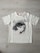 A white t-shirt with a printed graphic of a stylized cartoon girl's face, accompanied by bold black text saying 'ENJOY EVERY.' Below the graphic, the word 'MOMENT!' is repeated three times in light pink font. The shirt is laid flat on a light wooden surface.