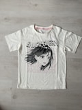 A white t-shirt with a printed graphic of a stylized cartoon girl's face, accompanied by bold black text saying 'ENJOY EVERY.' Below the graphic, the word 'MOMENT!' is repeated three times in light pink font. The shirt is laid flat on a light wooden surface.