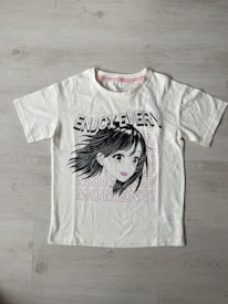 A white t-shirt with a printed graphic of a stylized cartoon girl's face, accompanied by bold black text saying 'ENJOY EVERY.' Below the graphic, the word 'MOMENT!' is repeated three times in light pink font. The shirt is laid flat on a light wooden surface.