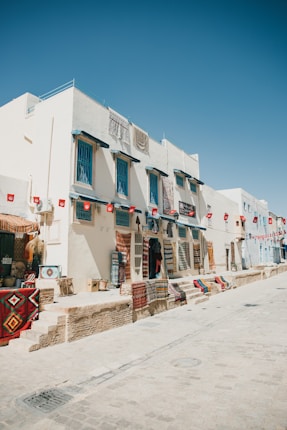 A sunlit street lined with shops featuring colorful textiles and carpets hanging from the walls. The buildings are painted in light colors with blue window frames, and small flags hang along the storefront. The sky is clear and bright, adding to the open and inviting atmosphere of the location.