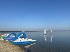 Paddle boats are lined up on a sandy beach with a peaceful, clear lake stretching into the distance. A set of white swings is installed on the water, and a clear blue sky meets the horizon.